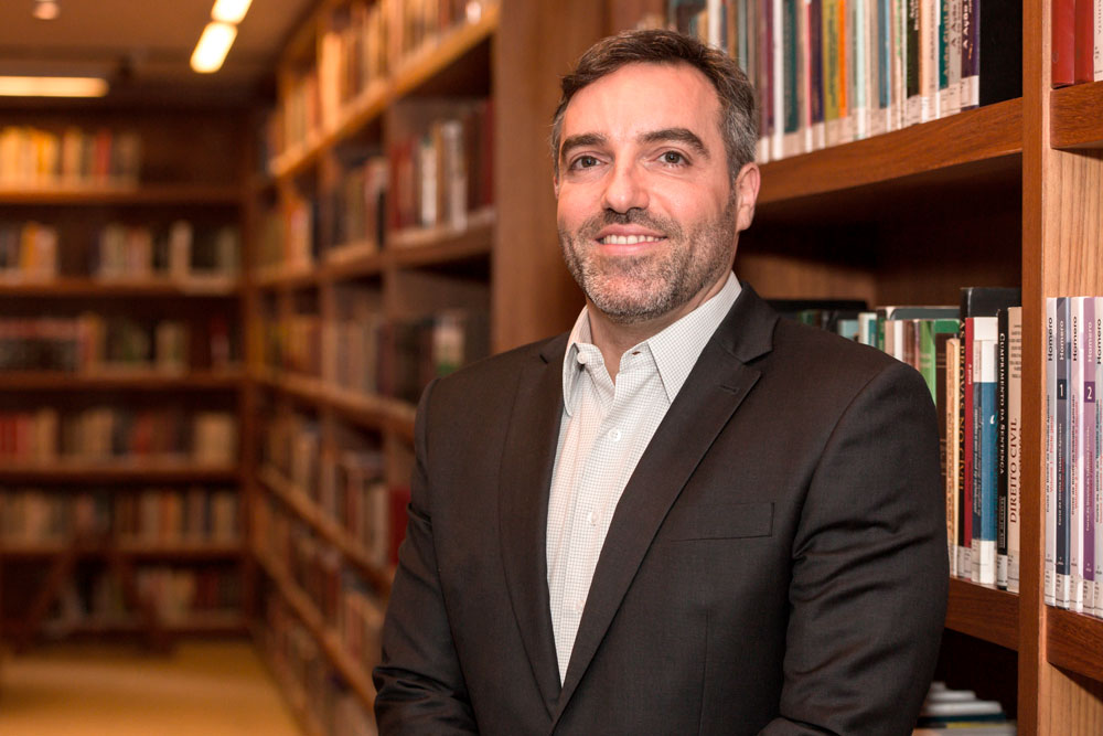 A picture of Fabio Ozi in a library. He is standing, smiling, wearing a white shirt and a dark gray suit. In the background, there are shelves with books.