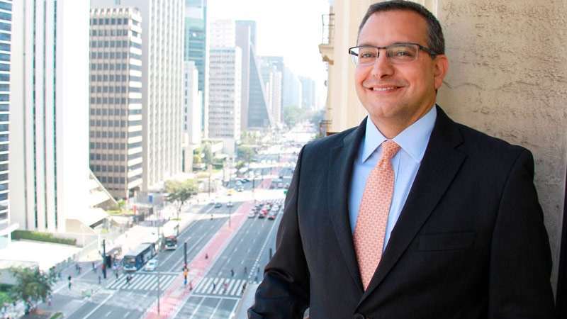 A picture of Marcelo Mansur, standing on a balcony. He is wearing a dark gray suit, light blue shirt, a tie printed with an orange and white geometric design and glasses. São Paulo's Avenida Paulista (Paulista Avenue) can be seen in the background.