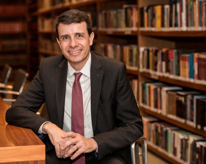 A picture of Paul Brancher in a library. He is sitting with his right arm leaning on a table and wearing a dark gray suit, light gray shirt, and wine-colored tie. In the background, there are shelves with books.
