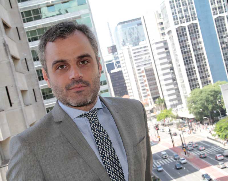 A picture of Rogério Taffarello, standing on a balcony. He is wearing a light gray suit, white shirt and tie with a black and white geometric print. São Paulo's Avenida Paulista (Paulista Avenue) can be seen in the background.